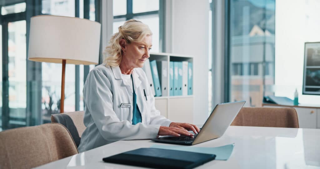 Doctor, laptop and medical with woman at desk in office of hospital for diagnosis, planning or research. Computer, healthcare and typing with mature medicine professional in clinic to update records.