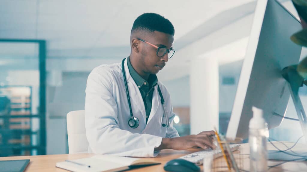 Black man, doctor and typing on computer for wellness, telehealth report or insurance records. Desktop, serious and medical worker with healthcare history, clinic schedule and research in office.