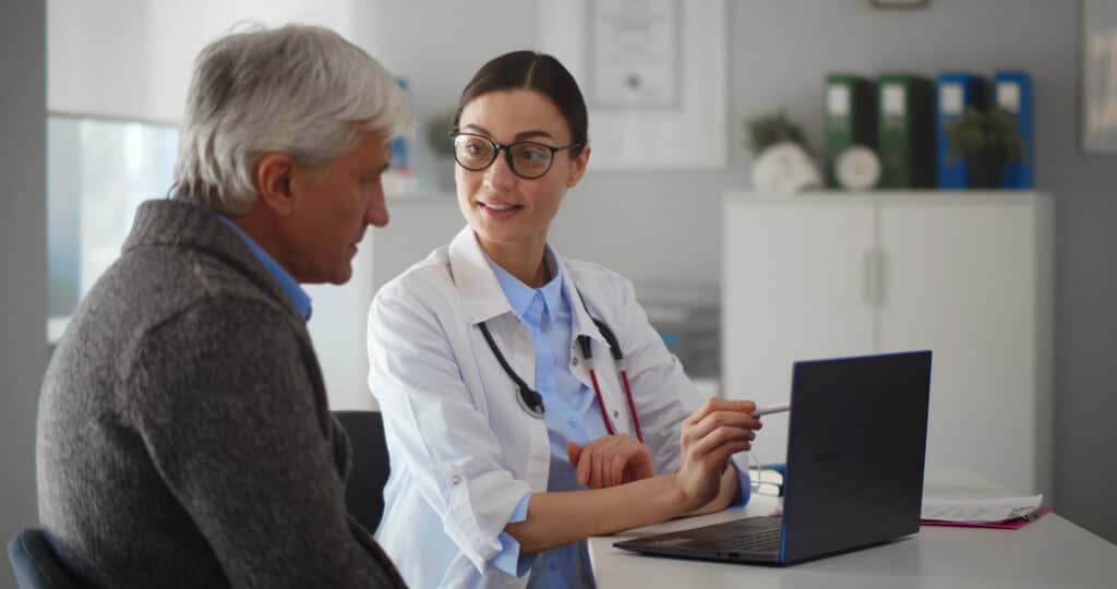 Young doctor talking to senior man patient sitting at desk in hospital office. Practitioner and patient looking at laptop screen discussing medical test results and diagnosis