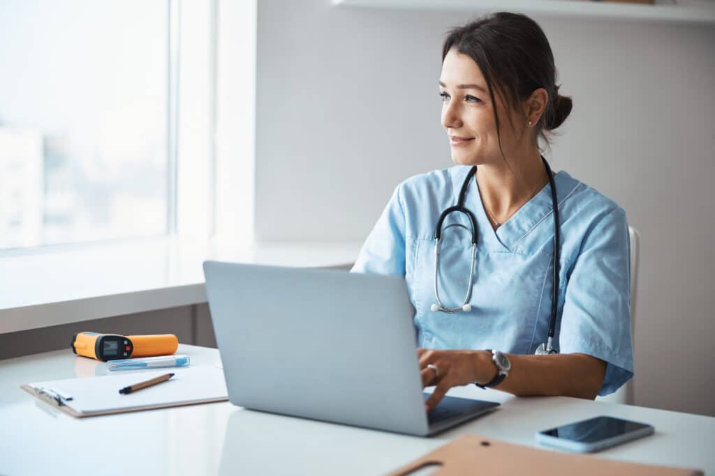 Beautiful young woman physician looking out the window and smiling while sitting at the table and working on notebook