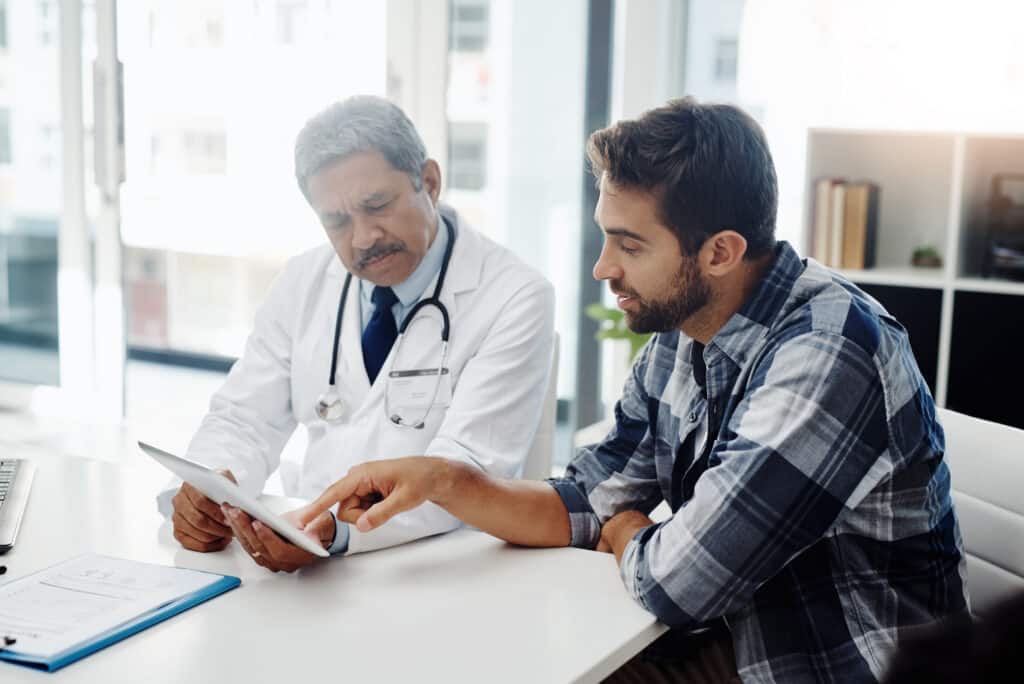 Healthcare, doctor and patient in consultation with tablet, expert advice, discussion and results. Medicine, health care and Indian man in doctors office, internet and consulting medical professional.