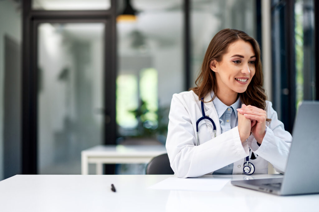 A happy adult pretty female doctor having an online video call on a laptop at the office.