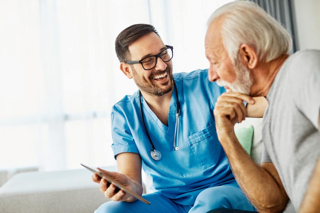 Doctor or nurse caregiver showing a tablet screen to senior man and laughing at home or nursing home